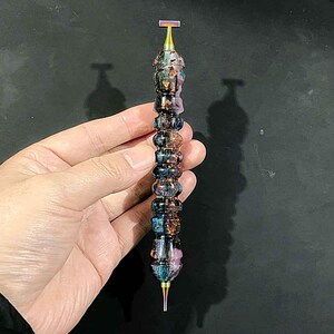 May include: A multi-faceted, cylindrical tool featuring a swirling, iridescent resin body with deep blue, purple, and copper-toned metallic flakes. The tool is capped with a metallic, rainbow-finished T-shaped top and a pointed, tapered tip at the base.