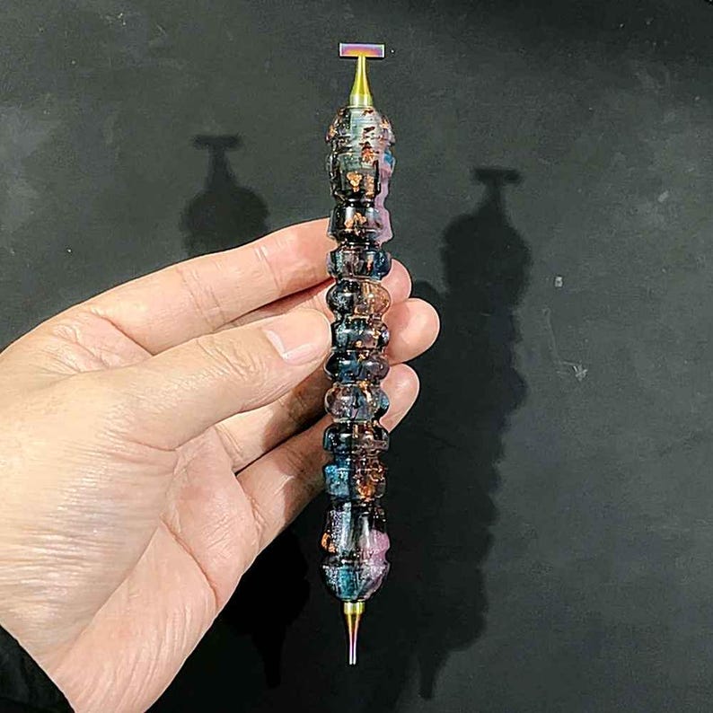 May include: A multi-faceted, cylindrical tool featuring a swirling, iridescent resin body with deep blue, purple, and copper-toned metallic flakes. The tool is capped with a metallic, rainbow-finished T-shaped top and a pointed, tapered tip at the base.