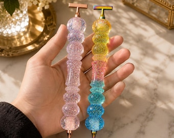 Könnte beinhalten: Zwei Stifte mit glitzernden, durchscheinenden, gestapelten kugelförmigen Körpern. Ein Stift hat einen hellrosa Farbton mit einer roségoldfarbenen T-förmigen Kappe und Spitze. Der andere Stift zeigt einen Farbverlauf in Gelb, Pink und Blau mit regenbogenfarbenem Metall.