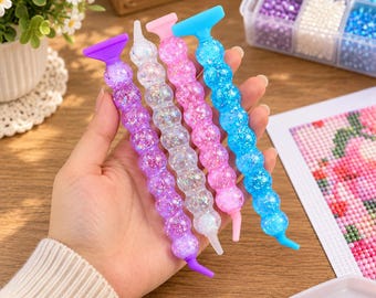 May include: Four diamond painting pens with textured, spherical, glitter-filled handles in purple, clear, pink, and blue. Each pen features a flat top and a pointed tip, designed for placing small beads onto a canvas for craft projects.