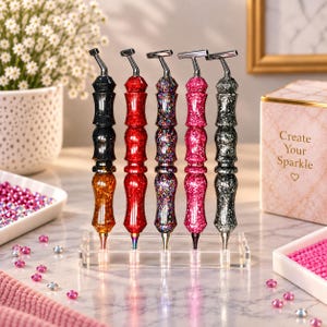 May include: Five glittery diamond painting pens with metallic T-shaped tips, arranged in a clear acrylic stand. The pens feature textured, multi-faceted barrels in black, red, multi-coloured, pink, and silver, each filled with sparkling glitter particles.