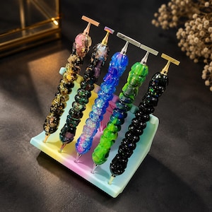 May include: Five diamond painting pens with textured, multi-colored resin handles featuring gold and silver-toned metal tips and T-shaped tops, arranged in a row on a pastel-colored, multi-slotted plastic stand designed to hold crafting tools.