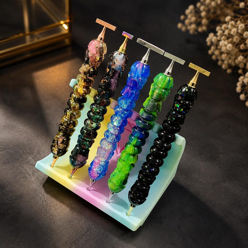 May include: Five diamond painting pens with textured, multi-colored resin handles featuring gold and silver-toned metal tips and T-shaped tops, arranged in a row on a pastel-colored, multi-slotted plastic stand designed to hold crafting tools.