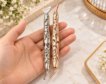 May include: Two metal diamond painting pens with intricate cut-out filigree patterns. One pen is finished in a polished silver tone, while the other features a rose gold tone. Both tools have a flat tip at the top and an angled tip at the bottom.