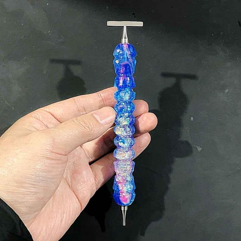 May include: A diamond painting drill pen featuring a textured, cylindrical body with a vibrant blue and iridescent finish. The tool includes a metallic silver T-shaped top and a pointed metal tip at the base, designed for precision crafting tasks.
