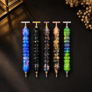 May include: Five diamond painting pens arranged vertically. Each pen features a textured, multi-segmented barrel with distinct color patterns: blue with pink flecks, black with iridescent glitter, dark multi-color, gold leaf, and green with blue swirls.
