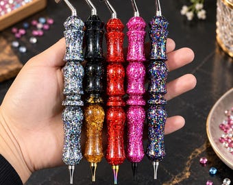 May include: Five diamond painting drill pens with glittery, multi-colored resin handles in silver, black, red, pink, and blue. Each pen features a contoured grip, a metallic top attachment, and a pointed metal tip for placing small craft beads.