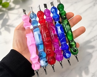 May include: Six diamond painting pens with contoured, translucent acrylic handles in pink, light blue, red, magenta, dark blue, and green. Each tool features a metallic tip at both ends, showcasing a marbled, swirling pattern within the material.