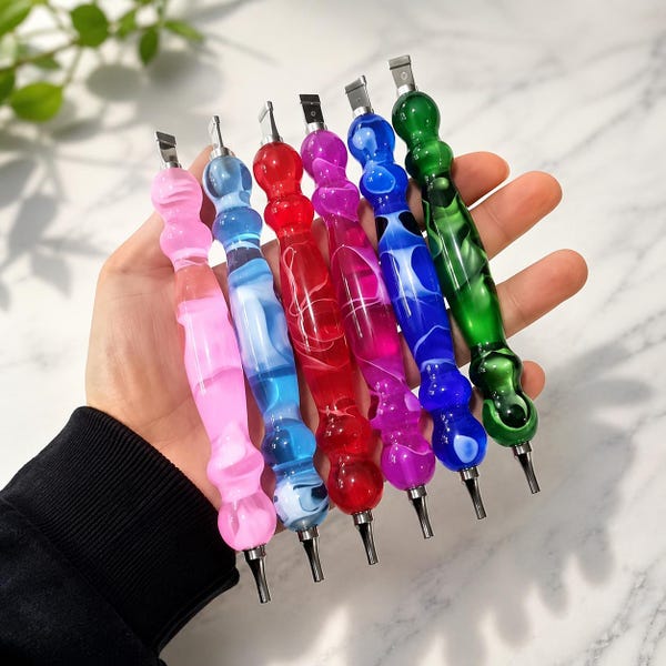 May include: Six diamond painting pens with contoured, translucent acrylic handles in pink, light blue, red, magenta, dark blue, and green. Each tool features a metallic tip at both ends, showcasing a marbled, swirling pattern within the material.