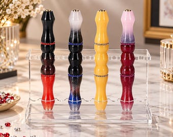 May include: Four decorative pens with contoured, multi-colored bodies stand upright in a clear acrylic display rack. Each pen features two bands of small, sparkling rhinestones. The pens display gradients of black, red, yellow, pink, and blue.