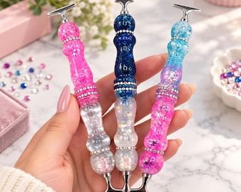 May include: Three diamond painting pens with T-shaped metal tips and silver rhinestone bands. The left pen is pink with glitter, the center pen is dark blue with glitter, and the right pen features a blue and pink gradient with glitter.