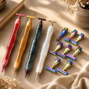 May include: Four diamond painting pens with glittery red, gold, dark blue, and white bodies, each featuring a metal T-shaped top. Eight interchangeable metal tips with a rainbow gradient finish are arranged to the right of the pens.