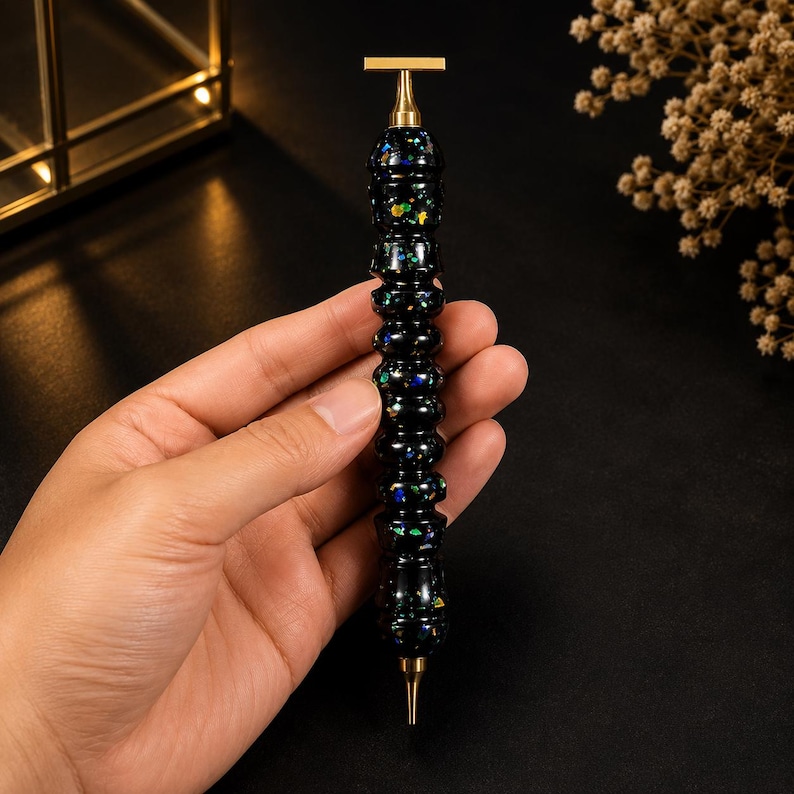 May include: A pen with a black barrel featuring embedded iridescent flecks of blue, green, and gold. The pen has a metallic gold-toned top with a horizontal bar and a matching gold-toned pointed tip at the bottom, with a segmented, ribbed body.