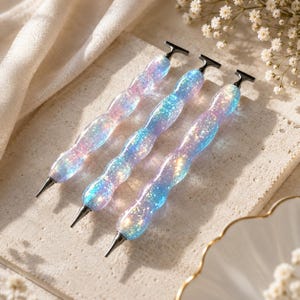 May include: Three diamond painting pens with wavy, ergonomic handles. Each pen features a translucent, iridescent body filled with shimmering blue, purple, and pink glitter particles. The pens have black metal tips and matching black T-shaped end caps.