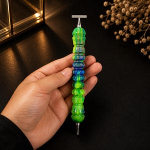 May include: A double-ended tool featuring a textured, cylindrical handle with a vibrant marbled pattern of bright green and deep blue swirls. The handle has a metallic finish with subtle glitter accents. It is capped with a silver-toned metal T-bar at one end and a pointed metal tip at the other end.
