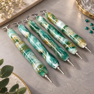Può includere: Cinque penne con clip argentate a forma di T e corpi caratterizzati da un motivo marmorizzato di verde smeraldo, bianco e scaglie di foglia d'oro. Ogni penna ha una punta argentata affusolata e una finitura liscia e lucida.