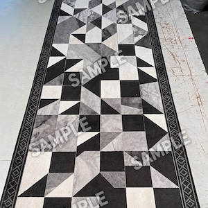 Op de afbeelding: Een lange rechthoekige loper met een geometrisch patroon van zwarte, witte en grijze driehoeken en vierkanten. Het kleed is omrand door een donkere lijst met een herhalend ruit- en kruispatroon. Het woord SAMPLE is herhaaldelijk afgedrukt.