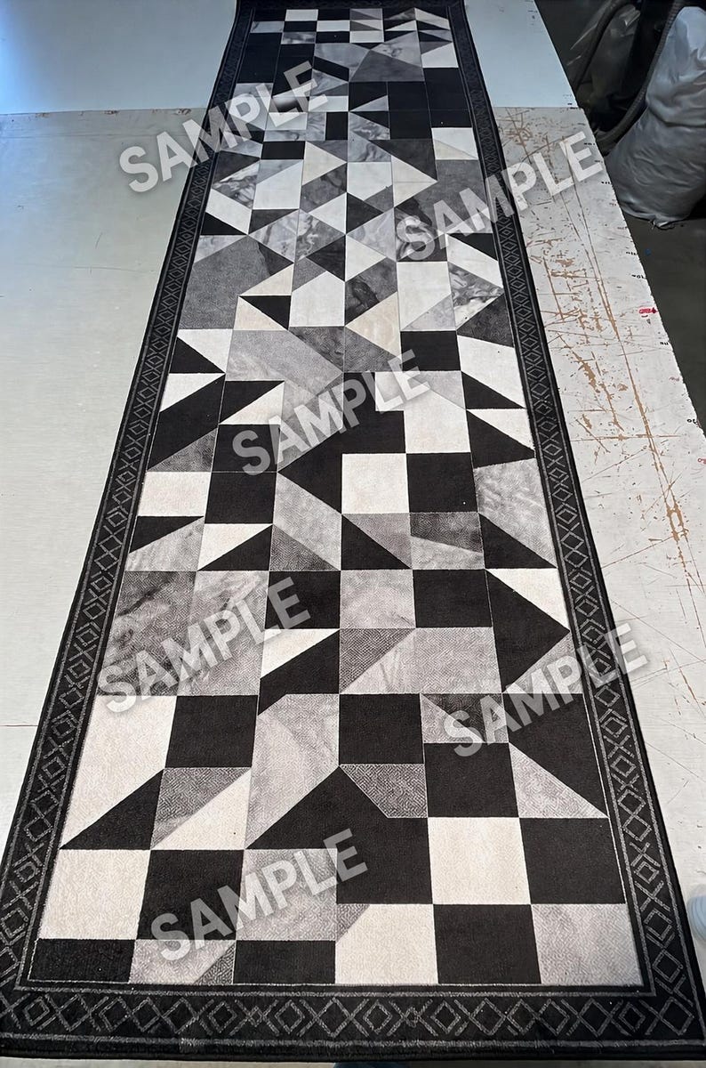 Op de afbeelding: Een lange rechthoekige loper met een geometrisch patroon van zwarte, witte en grijze driehoeken en vierkanten. Het kleed is omrand door een donkere lijst met een herhalend ruit- en kruispatroon. Het woord SAMPLE is herhaaldelijk afgedrukt.