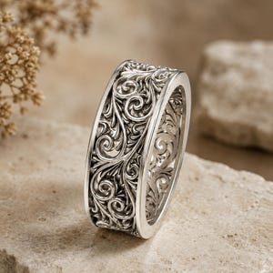 Puede incluir: Un anillo de metal plateado con un intrincado patrón en relieve continuo de enredaderas y hojas entrelazadas. La banda tiene un perfil ancho y plano con un acabado pulido, mostrando volutas detalladas que envuelven toda la superficie exterior.