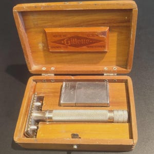 Peut inclure: Un rasoir de sûreté vintage Gillette et un étui à lames rectangulaire en métal rangés dans une boîte en bois. L'intérieur du couvercle présente un logo en forme de losange avec le texte Made in U.S.A. et Gillette Known the World Over.