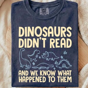 May include: A dark navy blue crew neck t-shirt featuring the light yellow text "DINOSAURS DIDN'T READ AND WE KNOW WHAT HAPPENED TO THEM" with light blue line art illustrations of four different dinosaurs positioned between the two lines of text.