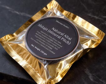 Può includere: Un contenitore rotondo di Shilajit (Mineral Pitch) Natural Altai di Siberian Healer, sigillato in una busta di plastica dorata. L'etichetta nera presenta testo bianco che dettaglia il nome del prodotto, gli ingredienti e un peso netto di 250 g.