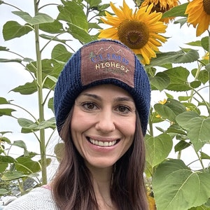 Pode incluir: Uma mulher usando um gorro azul com um remendo marrom que diz "California's Highest Peak" está em pé na frente de um campo de girassóis. Os girassóis estão em diferentes estágios de floração, alguns estão totalmente abertos e outros ainda fechados.