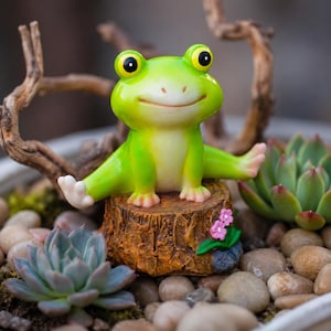 Puede incluir: Una figura de rana de color verde brillante con grandes ojos amarillos y una sonrisa sutil, sentada en posición de espagat sobre un tocón de árbol marrón texturizado. El tocón presenta un pequeño grupo de flores rosas con hojas verdes.