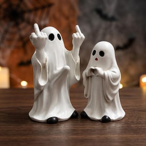 Peut inclure: Deux figurines de fantômes blanches en résine. Le plus grand fantôme à gauche a des yeux noirs et fait un geste avec les deux majeurs levés. Le plus petit fantôme à droite a des yeux noirs, une petite bouche rouge et forme un cœur avec ses mains.