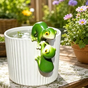 Könnte beinhalten: Ein weißer zylindrischer Übertopf mit vertikaler Rillenstruktur ist mit zwei grünen Froschfiguren am Rand verziert. Ein Frosch sitzt auf dem Rand und blickt in das Wasser, während der zweite Frosch vertikal darunter an der Außenwand hängt.