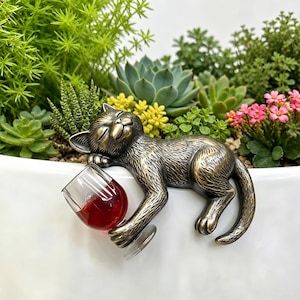 Könnte beinhalten: Eine bronzefarbene Metallkatzenfigur mit strukturierter Oberfläche, dargestellt in liegender Position mit geschlossenen Augen. Die Katze hält einen kleinen, durchsichtigen Glasbecher mit einer dunkelroten Flüssigkeit, der am Rand eines Gefäßes hängt.