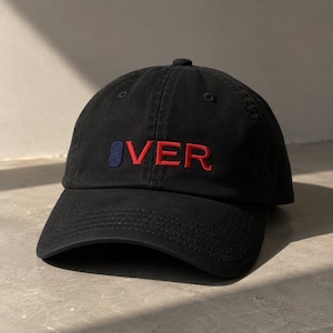 May include: A black baseball cap featuring an embroidered design on the front. The embroidery consists of a solid navy blue rectangle followed by the red text "VER" in a bold, sans-serif font. The cap has a curved brim and a structured crown.