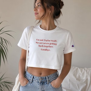 May include: A white short-sleeved crop top featuring red text that reads: I'm not Taylor Swift. We are never getting back together. Goodbye. The shirt has a small blue, red, and white Champion logo patch on the left sleeve and a crew neckline.