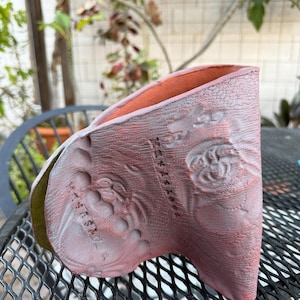 May include: A handmade, red-toned ceramic vase with a textured surface and embossed floral designs. The interior is a contrasting orange-red. The vase has a unique, asymmetrical shape and is displayed outdoors.