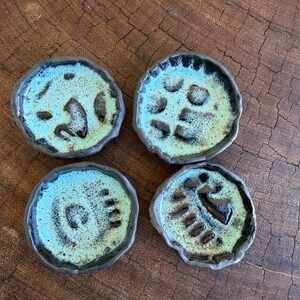 Puede incluir: Cuatro pequeños platos redondos de cerámica con un esmalte turquesa moteado y bordes marrón oscuro. Cada plato tiene un diseño abstracto único en marrón oscuro. Los platos están dispuestos sobre una superficie de madera oscura.