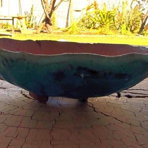 Pode incluir: Uma tigela de cerâmica artesanal com uma forma orgânica única. A tigela apresenta um exterior turquesa e um interior rosa-avermelhado. Assenta em três pequenos pés e é exibida numa superfície de madeira envelhecida.