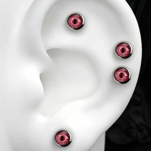 Può includere: Quattro orecchini a lobo circolari con un realistico design a occhio di drago rosa e rosso con una pupilla nera verticale. Ogni orecchino è incastonato in una lunetta in metallo color argento ed è esposto su un modello di orecchio bianco.