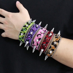 May include: A set of five wide bracelets featuring a leopard print pattern in neon green, purple, pink, red, and brown. Each bracelet is adorned with silver-toned metal spikes and circular colored gemstones that match the base color.