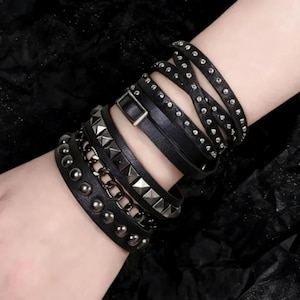 Op de afbeelding: Een meerlaagse zwarte armbandset met een combinatie van gladde leerachtige banden, zilverkleurige metalen piramidevormige studs, een donker metalen schakelkettinggedeelte en een rij ronde metalen studs, vastgezet met een rechthoekige gesp.