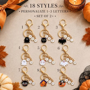 May include: A collection of nine gold-toned metal keychains featuring Halloween-themed enamel charms including ghosts, cats, and pumpkins. Each keychain includes a lobster clasp, a split ring, and small circular letter charms for personalization.