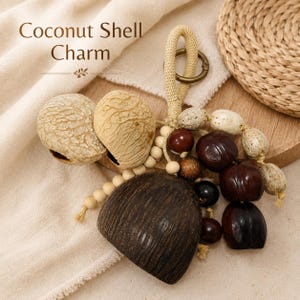 Peut inclure: Une breloque décorative composée d'un grand segment de coque de noix de coco brun foncé, de deux morceaux de coque plus clairs et texturés, et d'un groupe de perles polies brun foncé et blanc tacheté. Une boucle en corde beige avec un anneau.