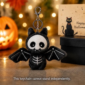 May include: A plush bat keychain featuring black faux fur, a white skull-shaped face with large black eyes, and white embroidered rib cage details on the chest. The wings have white stitching, and it includes a silver metal lobster clasp and ring.