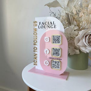 May include: A vertical acrylic sign featuring a white arched panel and a pink oval overlay. The sign displays the text FACIAL LOUNGE OKC, SCAN TO FOLLOW US, and Leave a review. It includes three social media icons with gold-bordered QR codes.