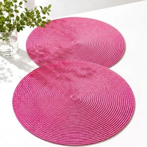 May include: Two circular hot pink placemats featuring a tightly coiled, braided texture. Each placemat displays a consistent spiral pattern radiating from the center to the outer edge, creating a uniform, flat surface for table settings.