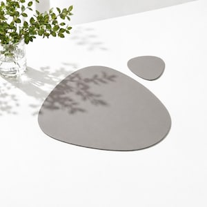 Peut inclure: Un ensemble de deux sets de table gris mat et lisses aux formes organiques irrégulières rappelant des galets. Le plus grand set est placé au centre, tandis que la plus petite pièce assortie est située en haut à droite, avec des bords arrondis.