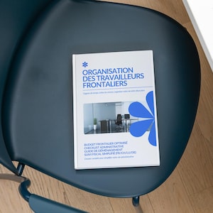 May include: A white book titled ORGANISATION DES TRAVAILLEURS FRONTALIERS with blue text and a blue flower graphic on the cover. The cover features a small photograph of an office interior. The book is resting on a dark blue chair.
