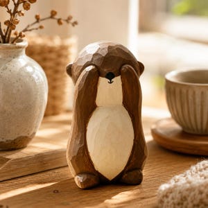 Könnte beinhalten: Eine handgeschnitzte Holzfigur eines Otters mit dunkelbraunem Kopf und Rücken, cremefarbenem Bauch und Pfoten, die vor das Gesicht gehalten werden. Die Oberfläche zeigt sichtbare Meißelspuren, eine kleine schwarze Nase und einen Mund.