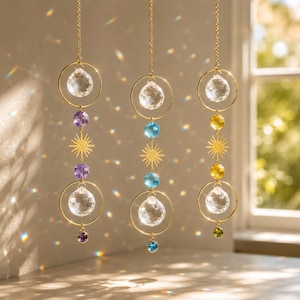 Könnte beinhalten: Drei hängende Sonnenfänger mit goldfarbenen Ketten und Ringen, klaren facettierten Kristallkugeln und zentralen sonnenförmigen Metallanhängern. Jedes Stück enthält kleinere farbige Kristallakzente in Violett, Blau und Gelb, die Licht brechen.