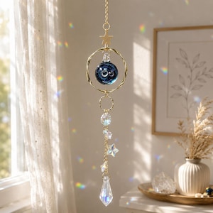 Op de afbeelding: Een hangende zonnevanger met een goudkleurige metalen ketting, een sterbedel, een gedraaid cirkelvormig frame en een donkerblauwe glazen bol. Onder de bol hangt een verticale ketting met heldere, gefacetteerde kristallen kralen in stervorm.
