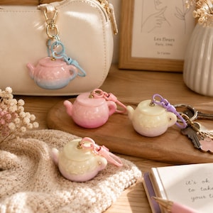 Könnte beinhalten: Vier Miniatur-Teekannen-Schlüsselanhänger in Pastelltönen wie Rosa, Blau und Creme. Jede Teekanne hat eine glänzende Oberfläche, einen kleinen Deckel und einen Henkel und ist an einem passenden Kunststoffverschluss befestigt. Einer ist an einer cremefarbenen Handtasche befestigt.